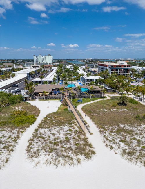 Aerial view of a beach town with white sand, a boardwalk, and low-rise buildings near turquoise water, under a partly cloudy blue sky.