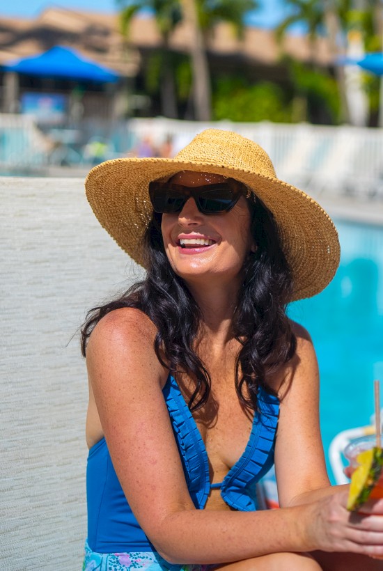 Two women relaxing by a pool in swimsuits, sun hats, and sunglasses; one holds a drink while the other waves, poolside lounge chairs nearby.