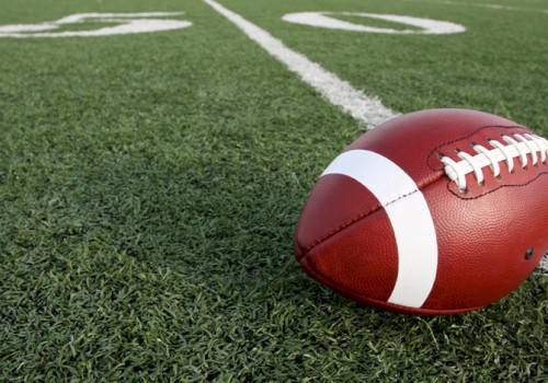 A close-up of a red football resting on a grassy football field near the white yard line, ready for play.