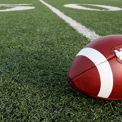 A close-up of a red football resting on a grassy football field near the white yard line, ready for play.