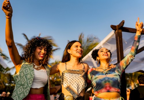 Three joyful friends celebrate outdoors, raising hands at a sunny festival with palm trees in the background.