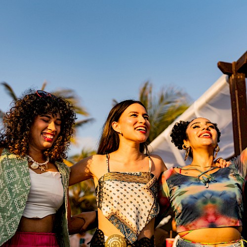 Three joyful friends celebrate outdoors, raising hands at a sunny festival with palm trees in the background.