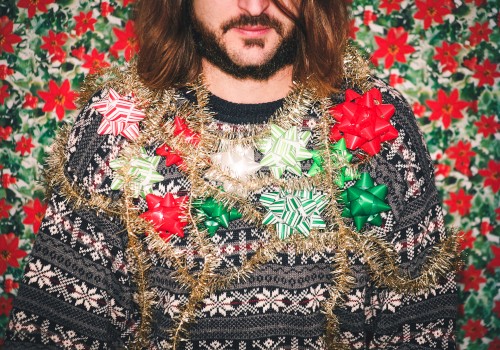 A bearded person in a patterned sweater adorned with shiny tinsel and oversized Christmas bows, against a festive poinsettia backdrop.