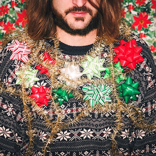A bearded person in a patterned sweater adorned with shiny tinsel and oversized Christmas bows, against a festive poinsettia backdrop.