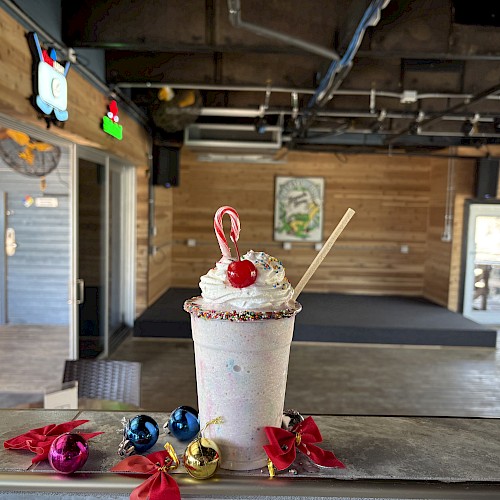 A tall milkshake with whipped cream, strawberry, and a stick on a wooden table, surrounded by colorful wrapped candies in a rustic shop.