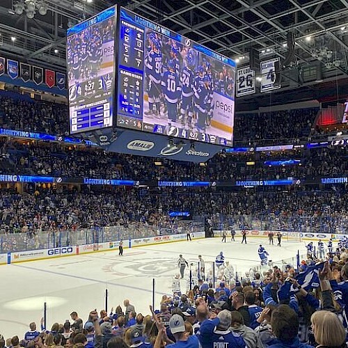 A crowded hockey arena during a game, fans on both sides, scoreboard above, ice rink glowing under bright lights.