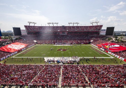 A packed football stadium with a green field, players on the pitch, and big red banners in the stands under a sunny sky.