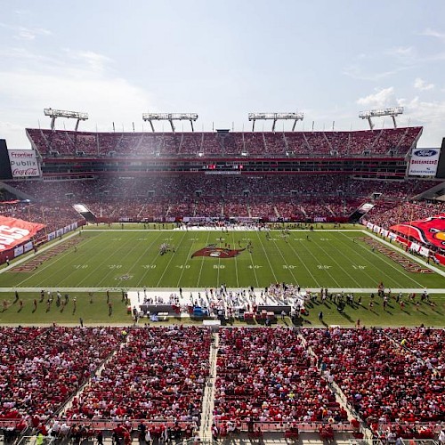 A packed football stadium with a green field, players on the pitch, and big red banners in the stands under a sunny sky.
