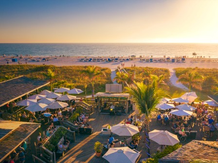 Beachside bar with palm trees, umbrellas, and crowds enjoying sunset near the ocean.