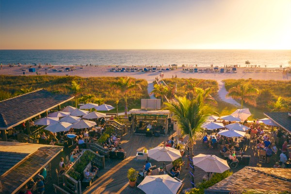 Beachside bar with palm trees, umbrellas, and crowds enjoying sunset near the ocean.