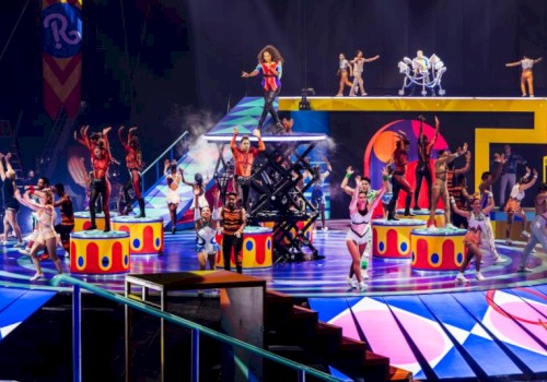 A colorful stage show with numerous performers in bright outfits, dancing on elevated platforms and stairs, surrounded by vibrant lights and props.