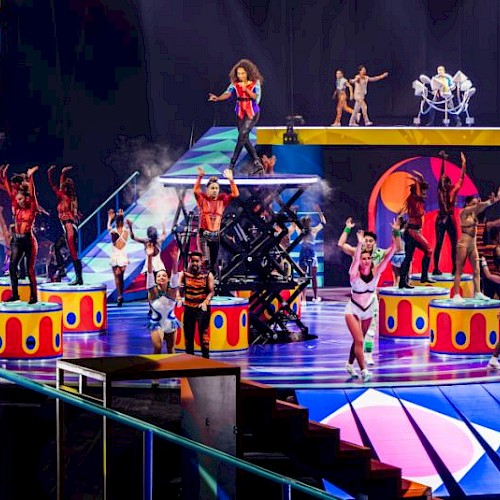 A colorful stage show with numerous performers in bright outfits, dancing on elevated platforms and stairs, surrounded by vibrant lights and props.
