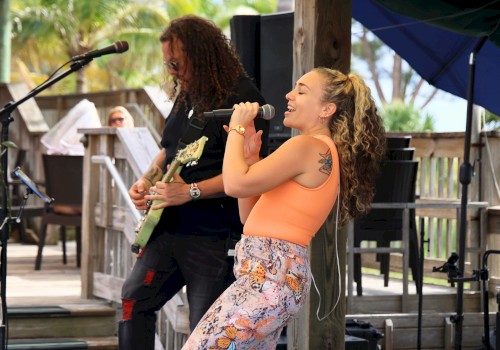 Two musicians perform on a sunny outdoor stage: a female singer in an orange top and a guitarist in the background, under a shaded canopy.