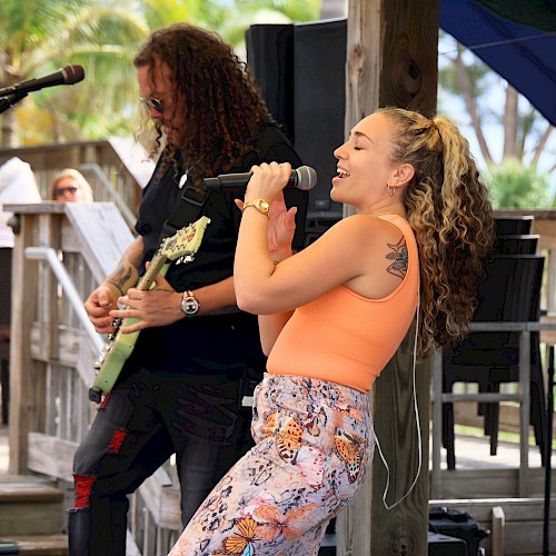 Two musicians perform on a sunny outdoor stage: a female singer in an orange top and a guitarist in the background, under a shaded canopy.