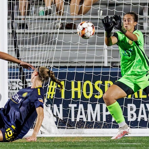 A goalkeeper in neon green saves a shot from a forward, while a defender in gray-blue and a teammate in navy react near the goal.