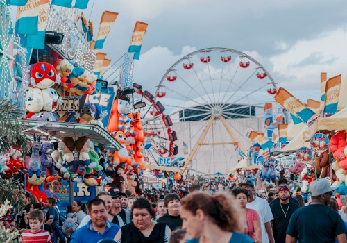 A crowded carnival scene with rides, a ferris wheel, colorful stalls, and people walking amid tents and balloons, under a cloudy sky.