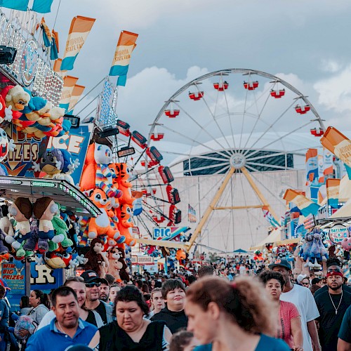 A crowded carnival scene with rides, a ferris wheel, colorful stalls, and people walking amid tents and balloons, under a cloudy sky.