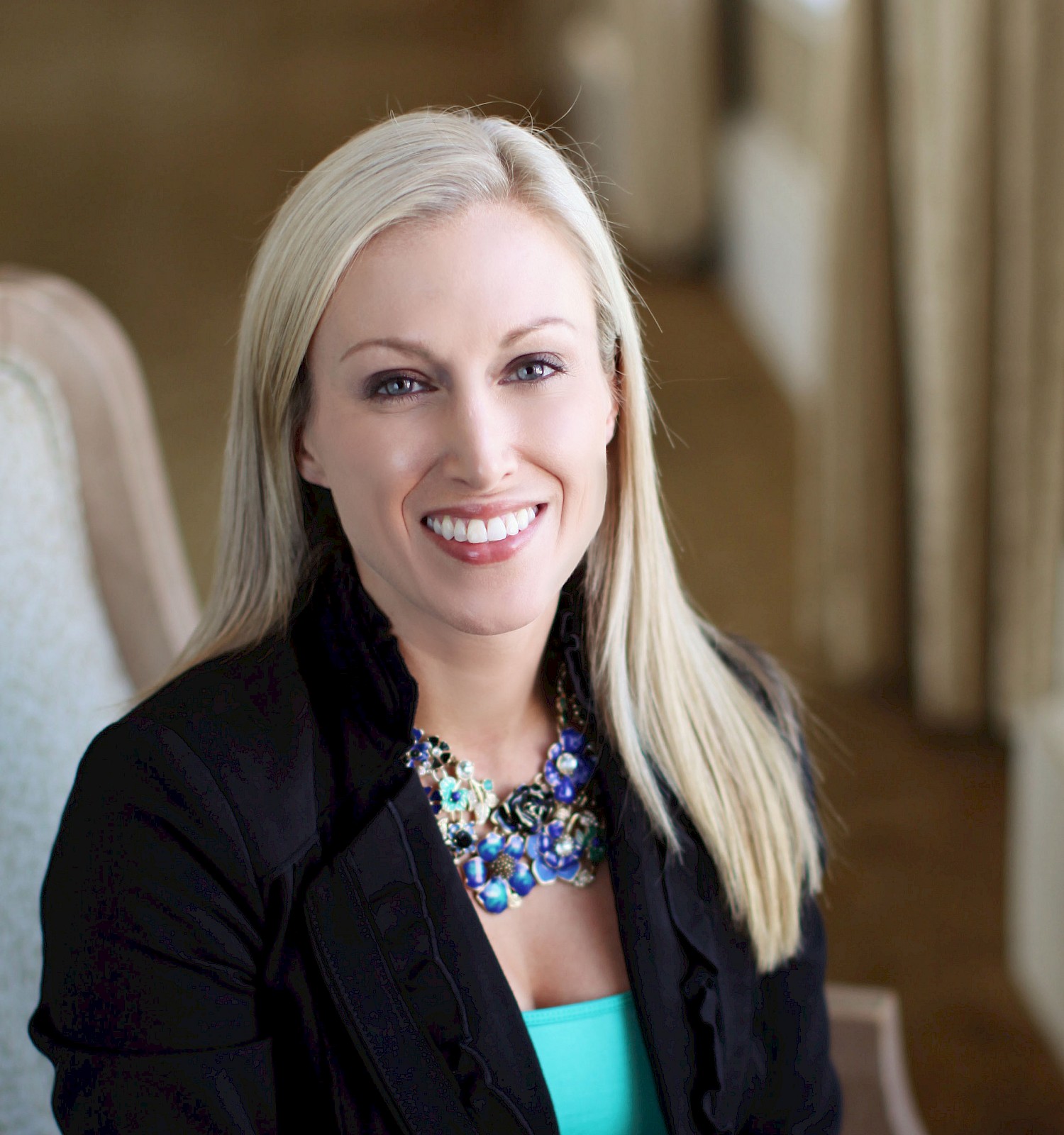 A smiling blonde woman in a black blazer and turquoise top sits in a light-colored chair, posing confidently in a well-lit indoor setting.