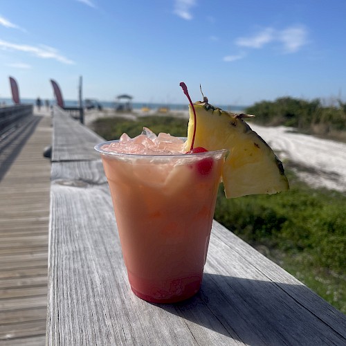 A pink grapefruit cocktail with ice, a pineapple slice garnish, and a cherry, set on a wooden railing by a sunny beach boardwalk.