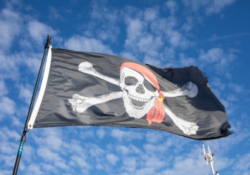 A pirate flag with a skull and crossbones flies against a blue sky, tattered edges and a red bandana waving.