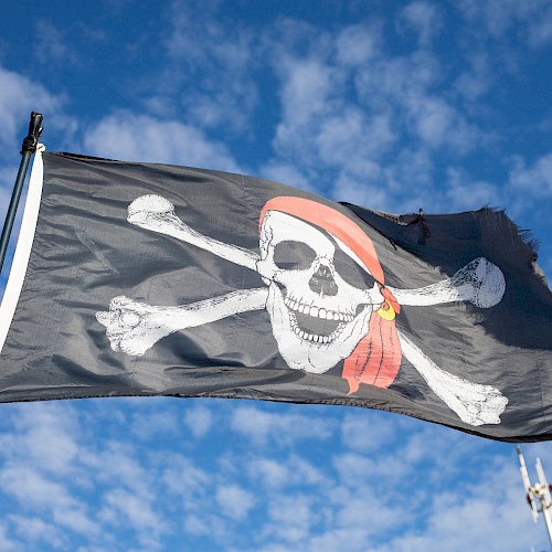 A Pirate flag with a skull and crossbones waves against a blue sky, a red bandana tied around the skull.