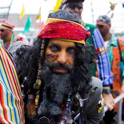 A bearded man in a pirate-like costume with a red bandana and colorful accessories, among others in festive outfits at a crowd.