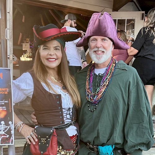 Two people in pirate/medieval costumes pose on a porch: woman in teal skirt, corset, hat; man in green tunic, purple hat, beads, belt bag. Ending.