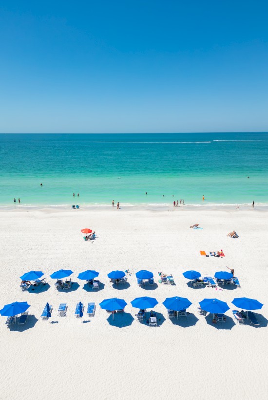 People enjoy a sunny beach with turquoise water, white sand, and many blue umbrellas lined up along the shore, while a few beachgoers swim nearby.