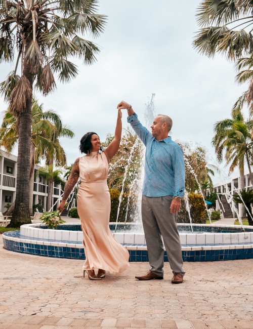 An older couple dances happily by a fountain, palm trees framing the scene, as they hold hands and twirl under a sunny sky.