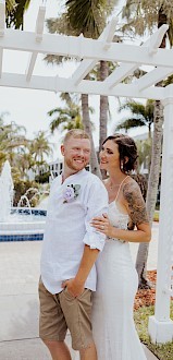 A couple posing for a beach wedding photo under a white arbor, with the groom in a white shirt and tan shorts and the bride in a white dress smiling together. End sentence.
