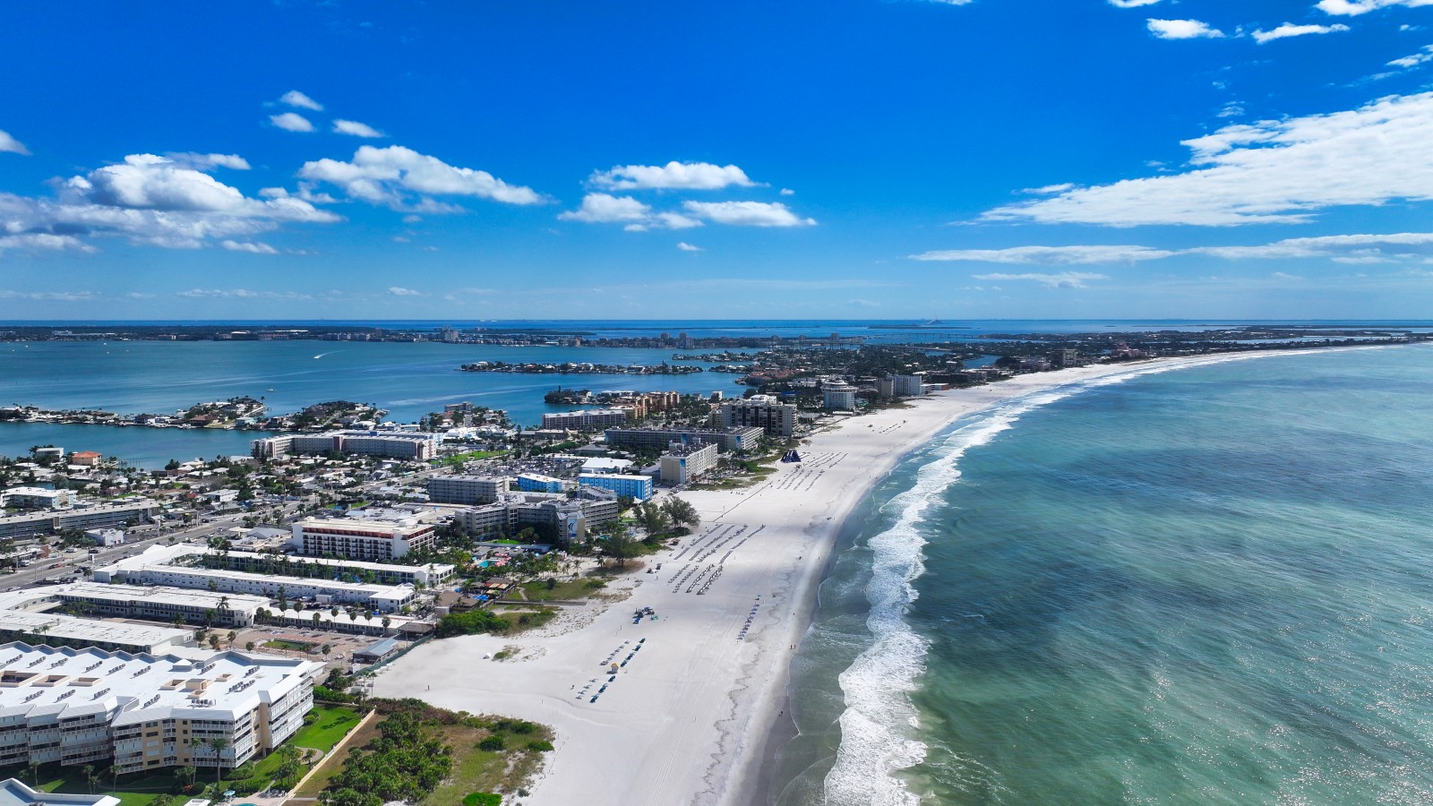 An aerial view of a coastal city with white sandy beaches, turquoise water, and numerous buildings under a partly cloudy sky.