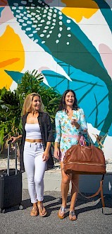 Two women are smiling and walking with luggage in front of a colorful, abstract mural and lush green plants.